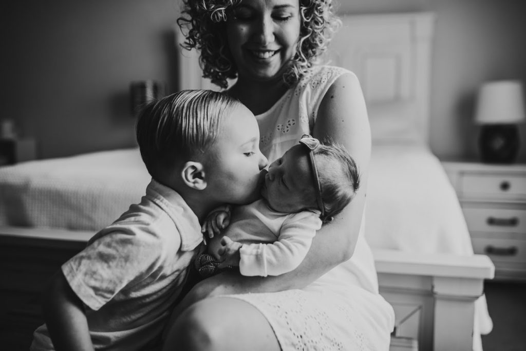 a happy mother holds her infant as her toddler kisses the infant